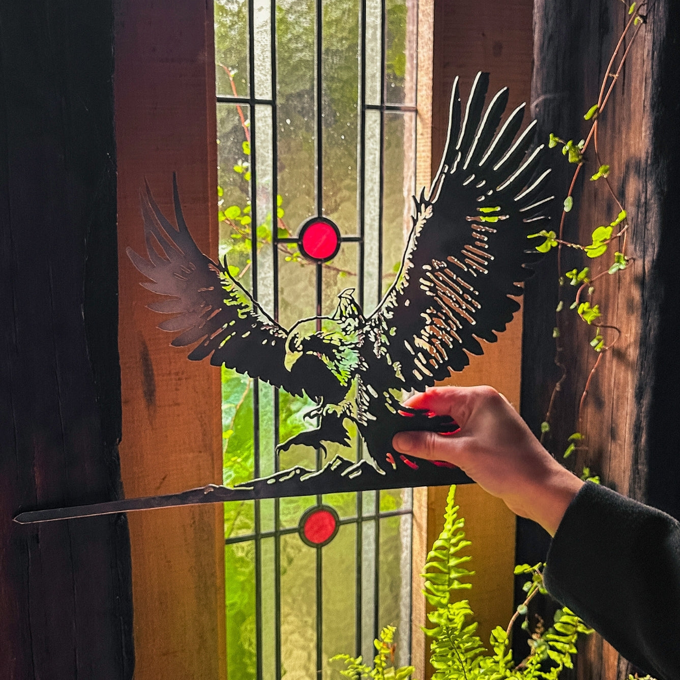 A hand holds the GANDALF™ ON GWAIHIR™ corten steel silhouette by Metalbird UK before a stained glass window with red circular accents, framed by wooden panels and lush green plants.