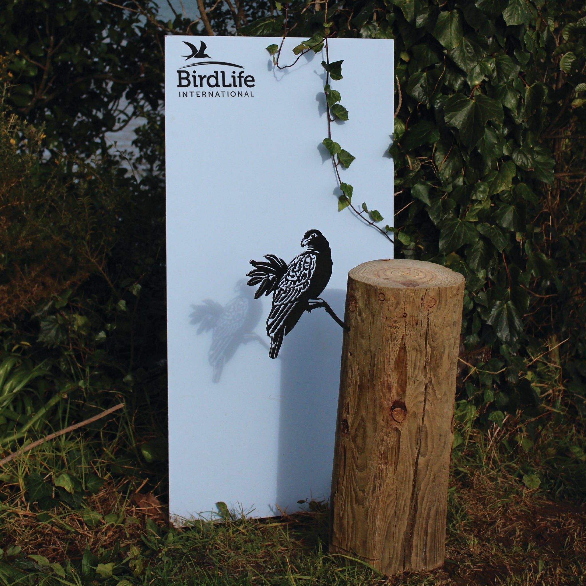The Manumea sign by Metalbird UK - CC features a black bird silhouette, displayed outdoors among greenery with a leafy vine overhead and a wooden stump in front.