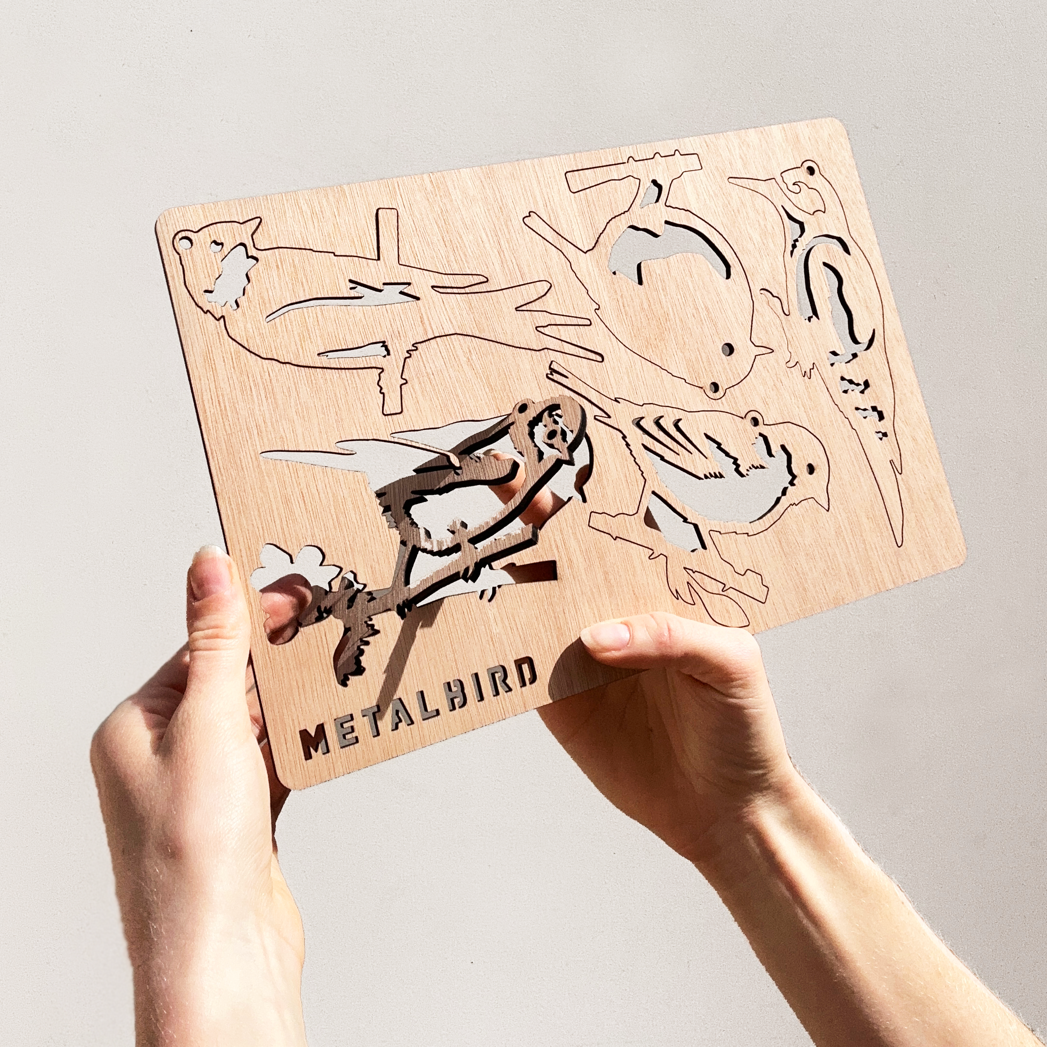 Two hands hold a thin Wooden Hanging Decoration by Metalbird UK, featuring bird silhouettes and the brand name at the bottom. Sunlight creates striking shadows, making it a unique gift for nature lovers.