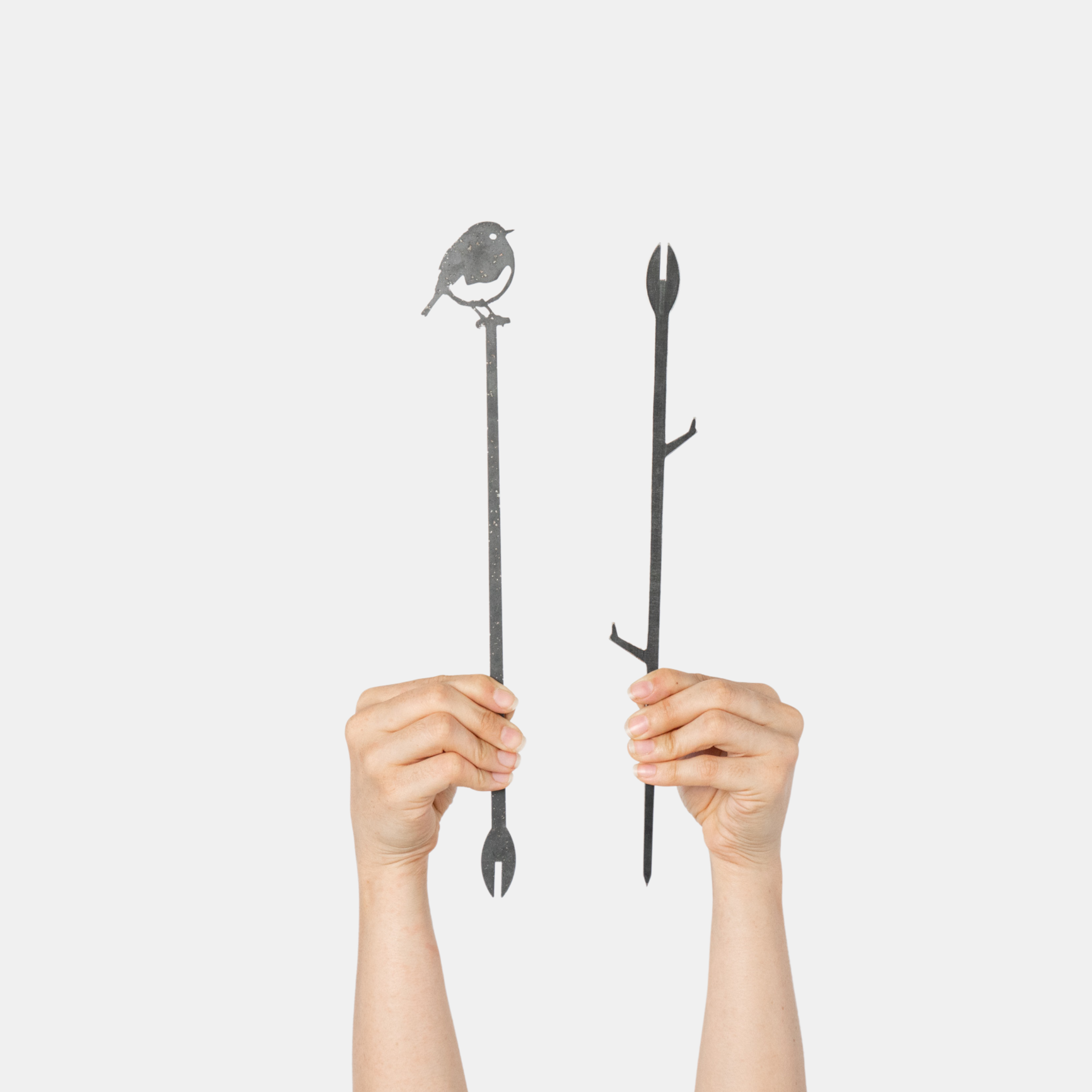 Two hands hold up the Pair Of Plant Stakes by Metalbird UK: one with a perched bird on top, the other with branch-like details—unique yard art accents—shown against a plain white background.