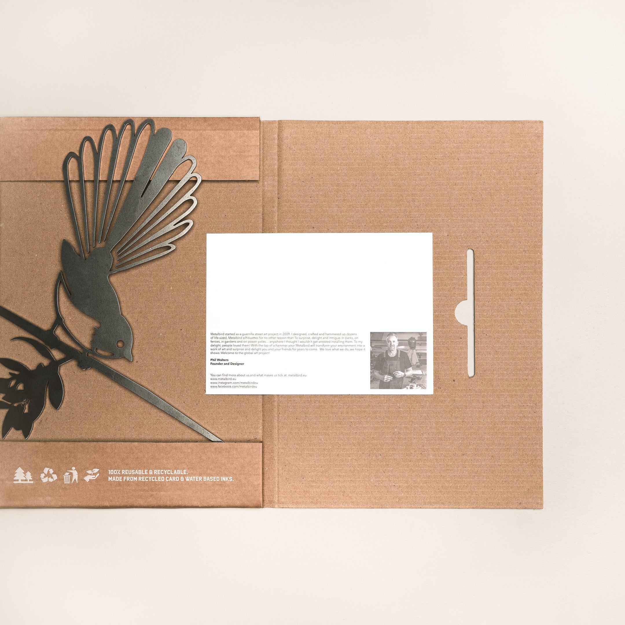 The Fantail (Piwakawaka) by Metalbird UK is a cardboard folder featuring a metal bird on a branch, reminiscent of yard art. A white card with text and photo peeks out on the right, set against a neutral background.