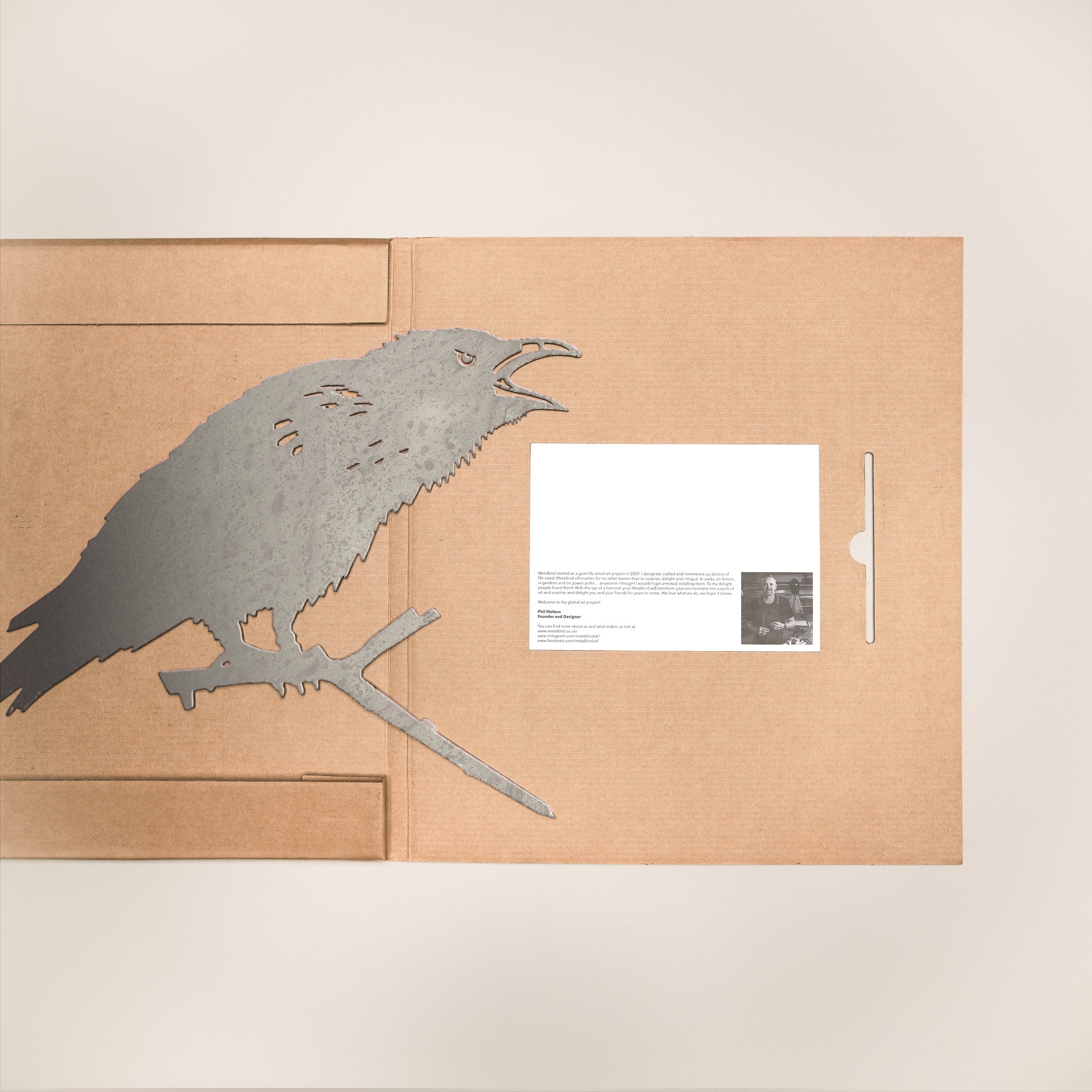 The Metalbird UK "Raven" features a metal garden art raven silhouette on a cardboard folder's left side, with a white card displaying printed text and a small black-and-white photo on the right.
