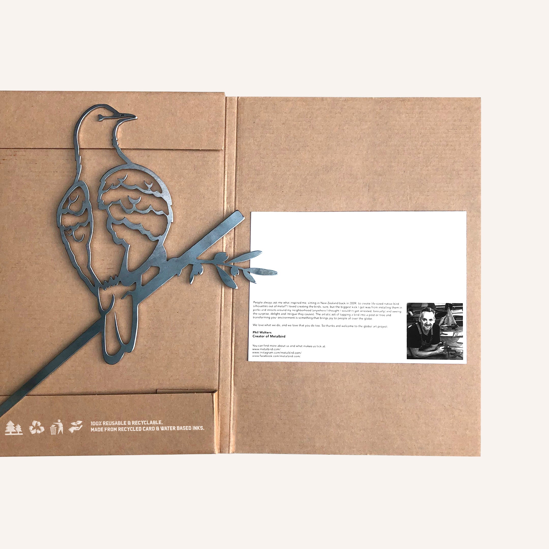 The Turtle Dove by Metalbird UK is a metal bird silhouette sculpture perched on a branch, displayed atop a brown cardboard folder with recycling icons, a printed message, and a black-and-white photo inside.