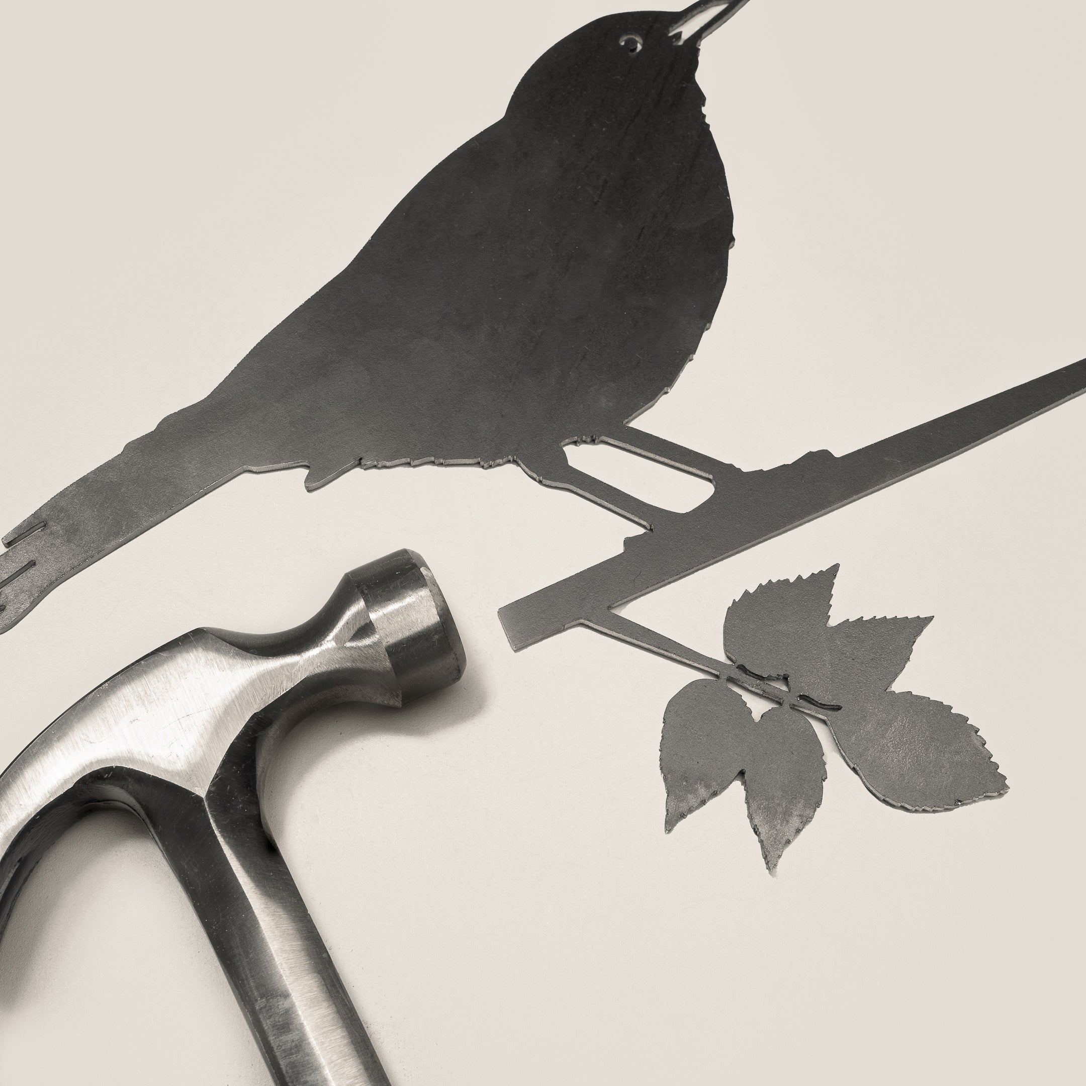 The Blackbird by Metalbird UK features a metal bird silhouette perched on a leafy branch beside a hammer, making unique yard art or a thoughtful gift, all shown against a light background.