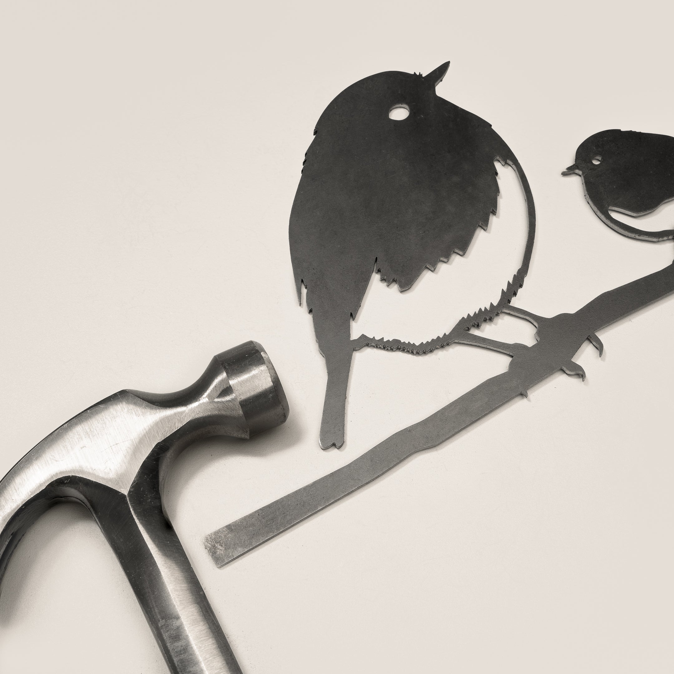 A metal hammer lies next to Metalbird UK's "Robin and Chick" corten steel cutouts, shaped like two birds on a branch—perfect for unique yard art—against a plain, light background.