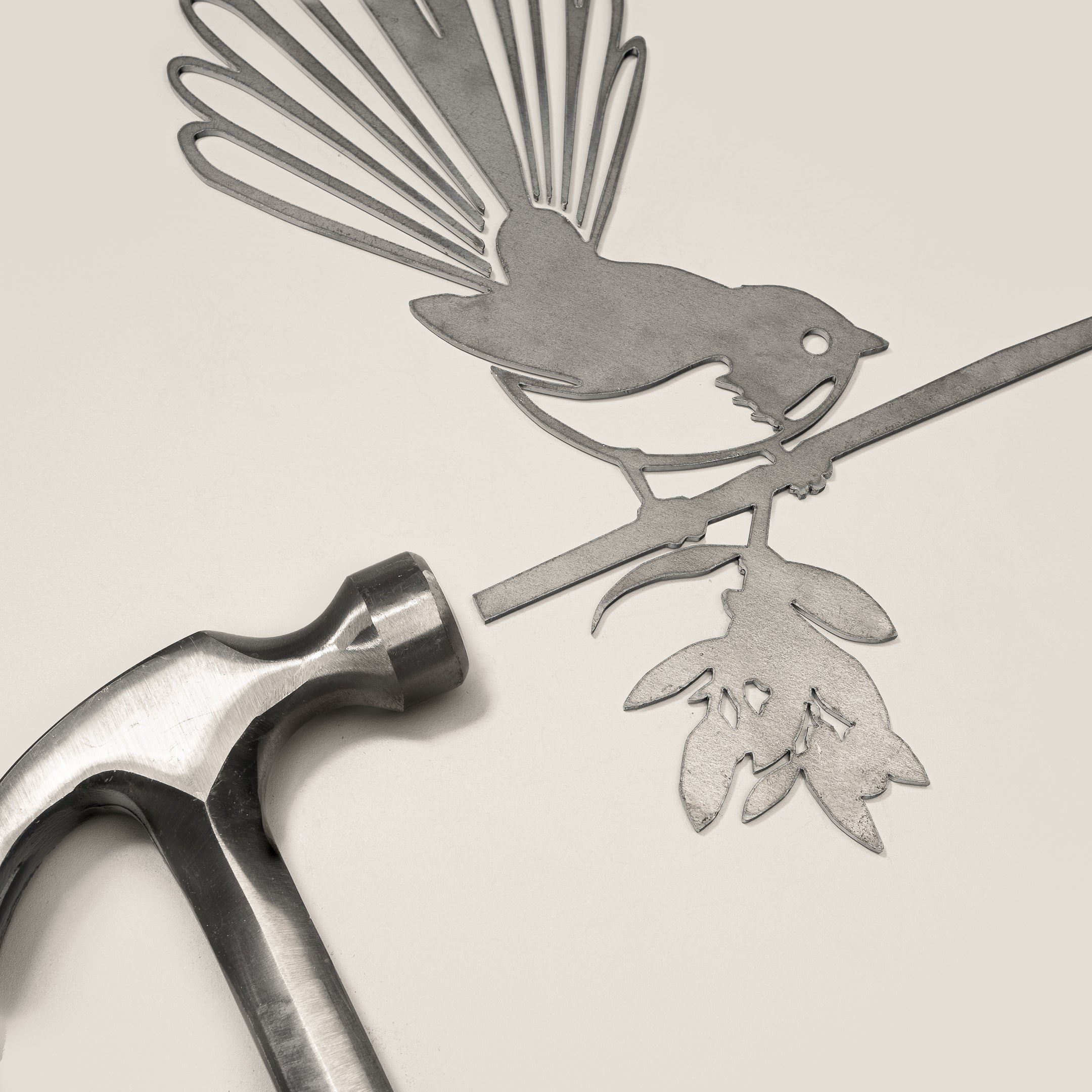 A metal hammer sits next to the Metalbird UK Fantail (Piwakawaka), a flat corten steel bird silhouette perched on a leafy branch—an elegant piece of garden art showcased on a light surface.