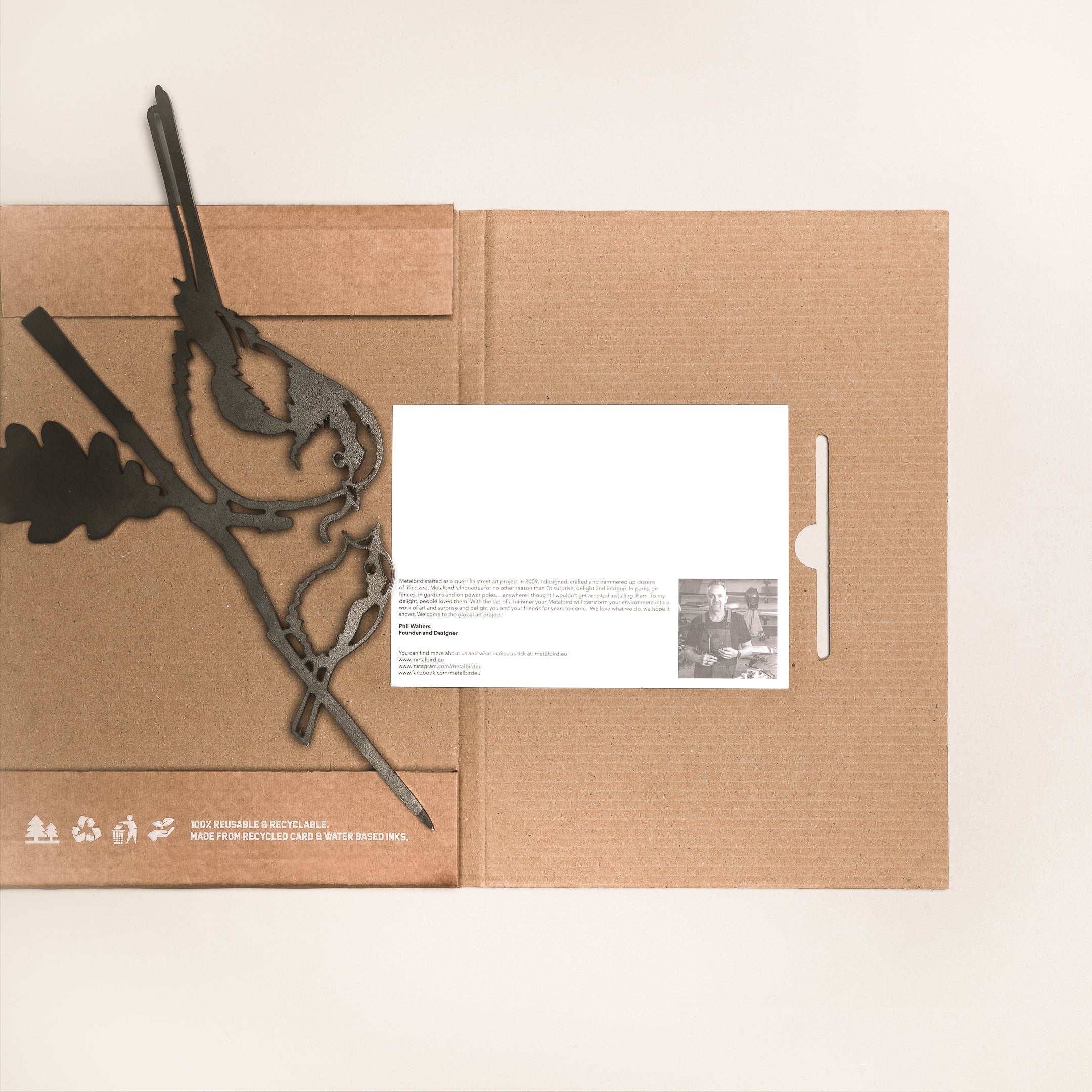 A flat-lay of Metalbird UK's "Long-tailed Tit & Chick" brown cardboard envelope, featuring a metal bird and leaf ornament—ideal for garden art or gifting—alongside a black pen and eco-friendly white card with printed text and photo.