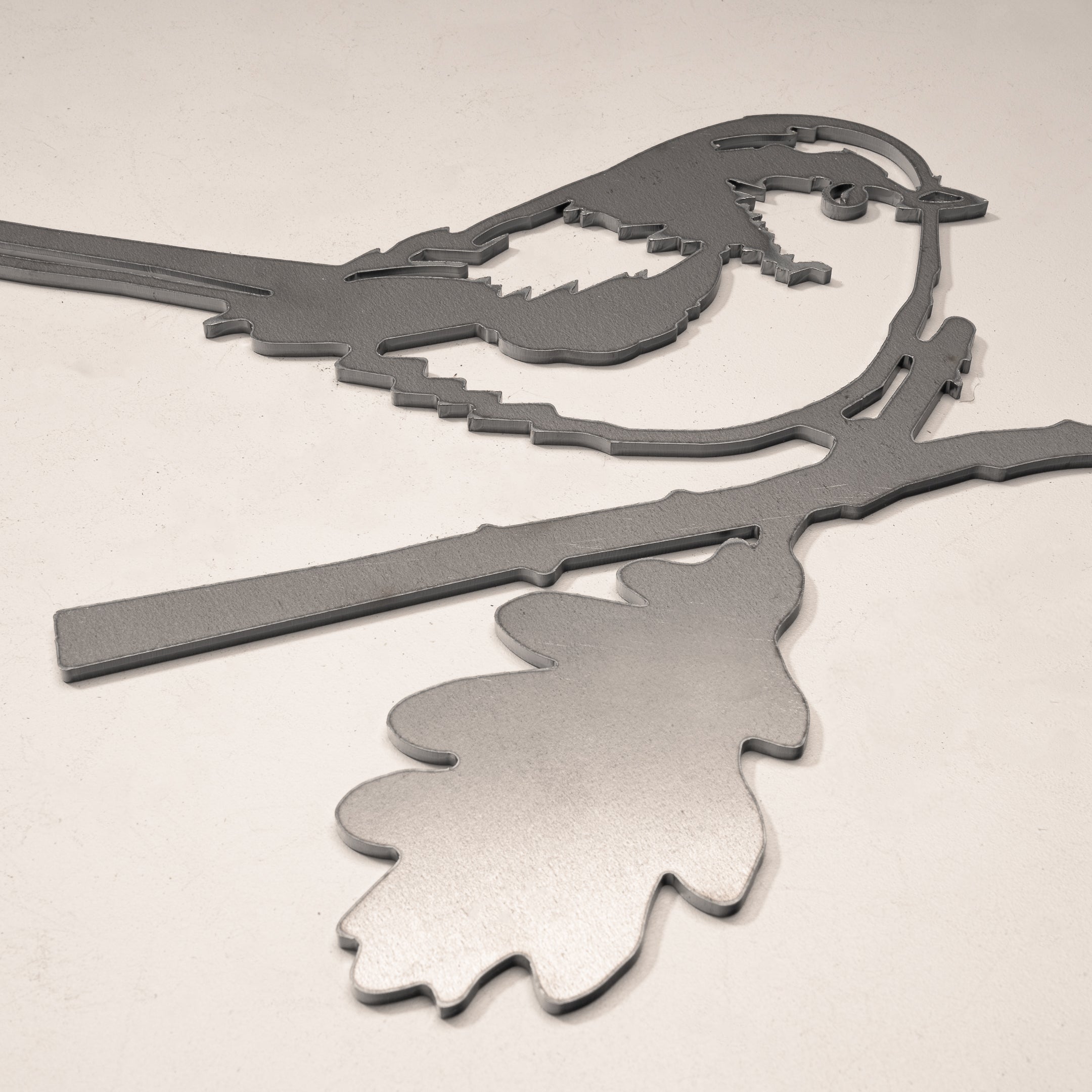 The Long-tailed Tit by Metalbird UK is a metallic, geometric silhouette of a bird on a branch with a large leaf, crafted from corten steel. Its clean lines and striking design make it standout yard art for any light-colored surface.