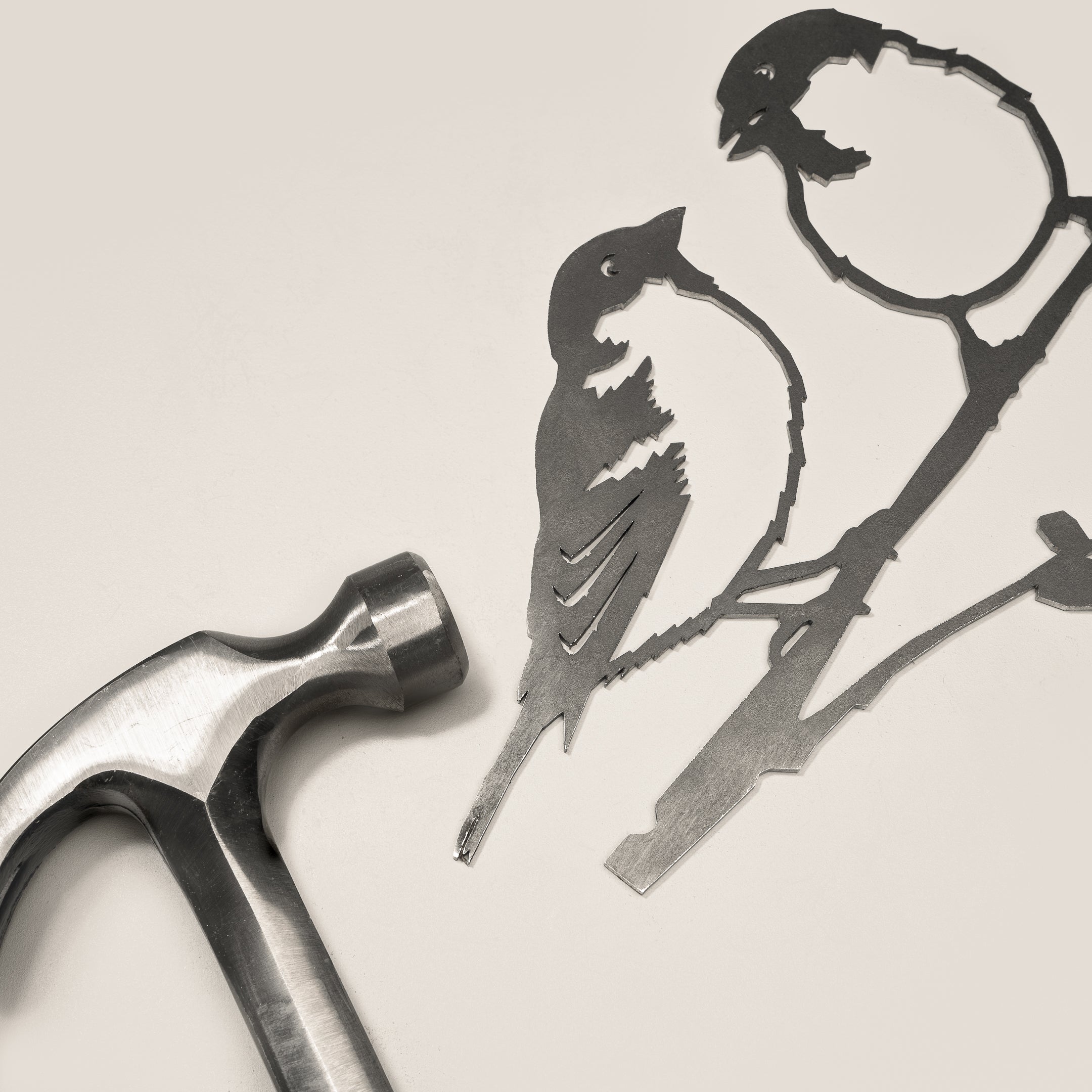 A metal hammer rests beside Metalbird UK's Pair of Sparrows, a delicate corten steel silhouette of two birds on branches, set against a light, neutral background.