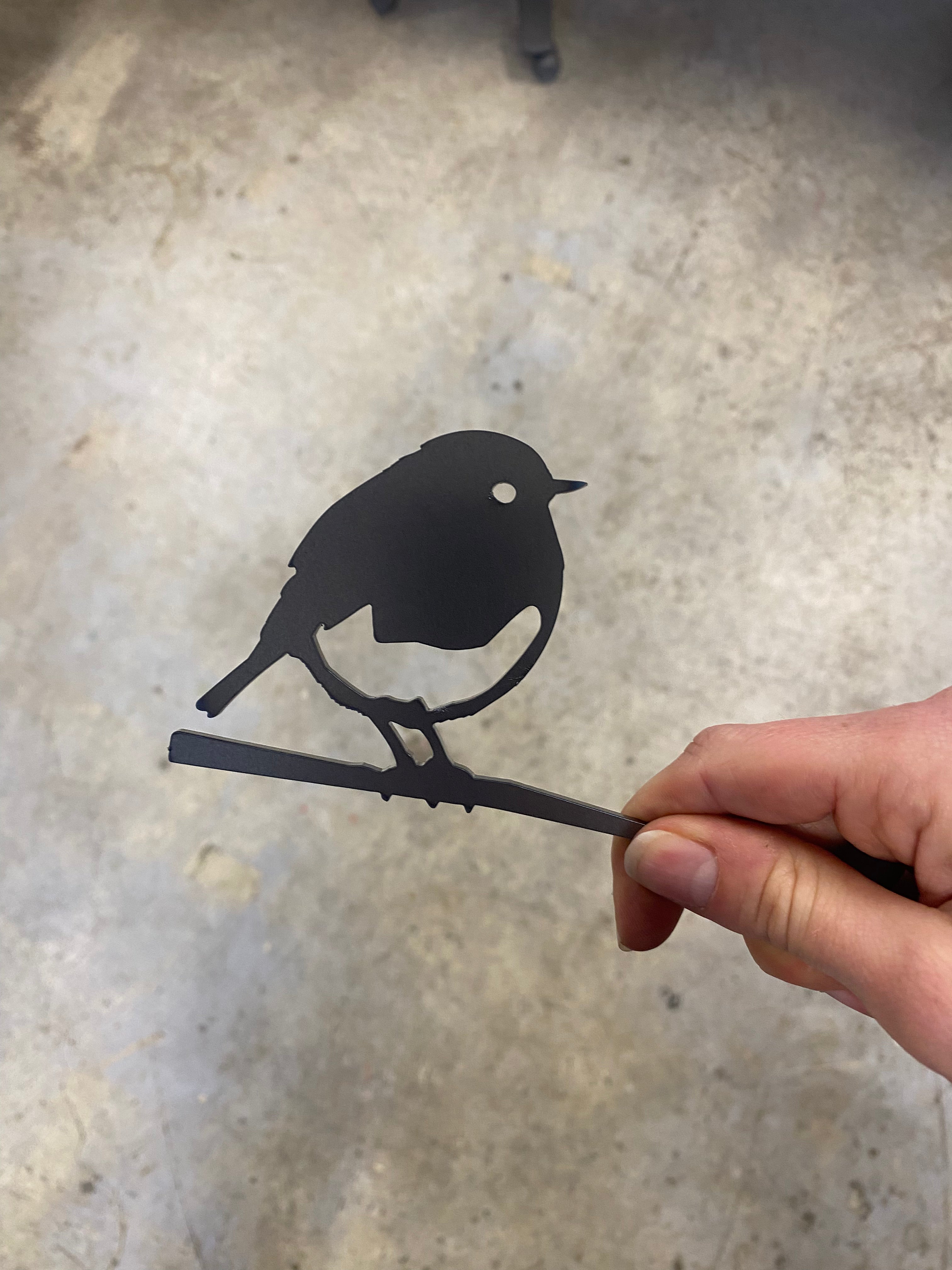 A hand holds Metalbird UK's FLOCK FAVOURITES, a small black metal bird silhouette on a branch—perfect garden art or thoughtful gift—shown against a blurred concrete background.