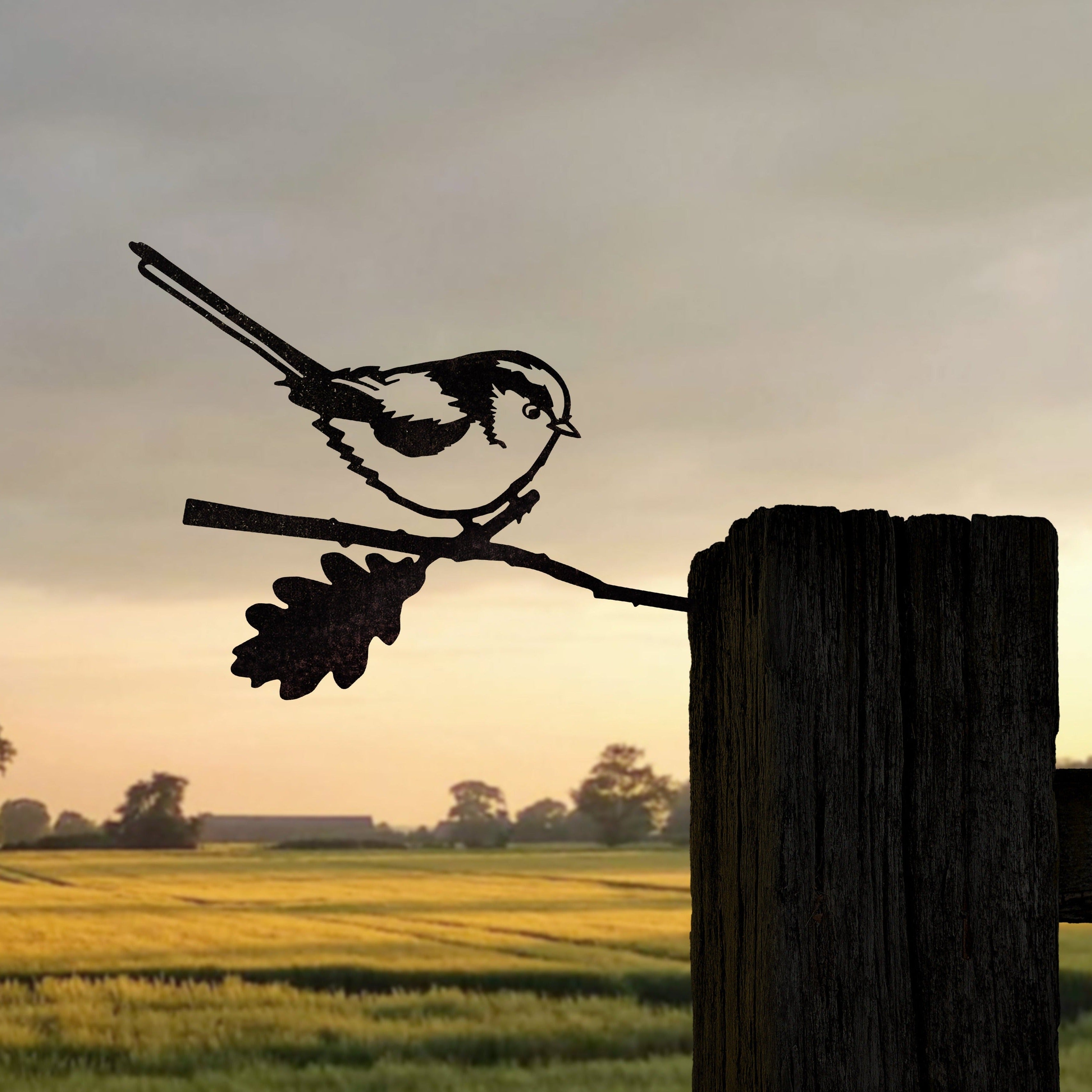 The Long-tailed Tit by Metalbird UK is a striking metal yard art silhouette perched on a branch and mounted on a wooden post—an ideal unique gift, beautifully highlighted against fields and trees at sunset.