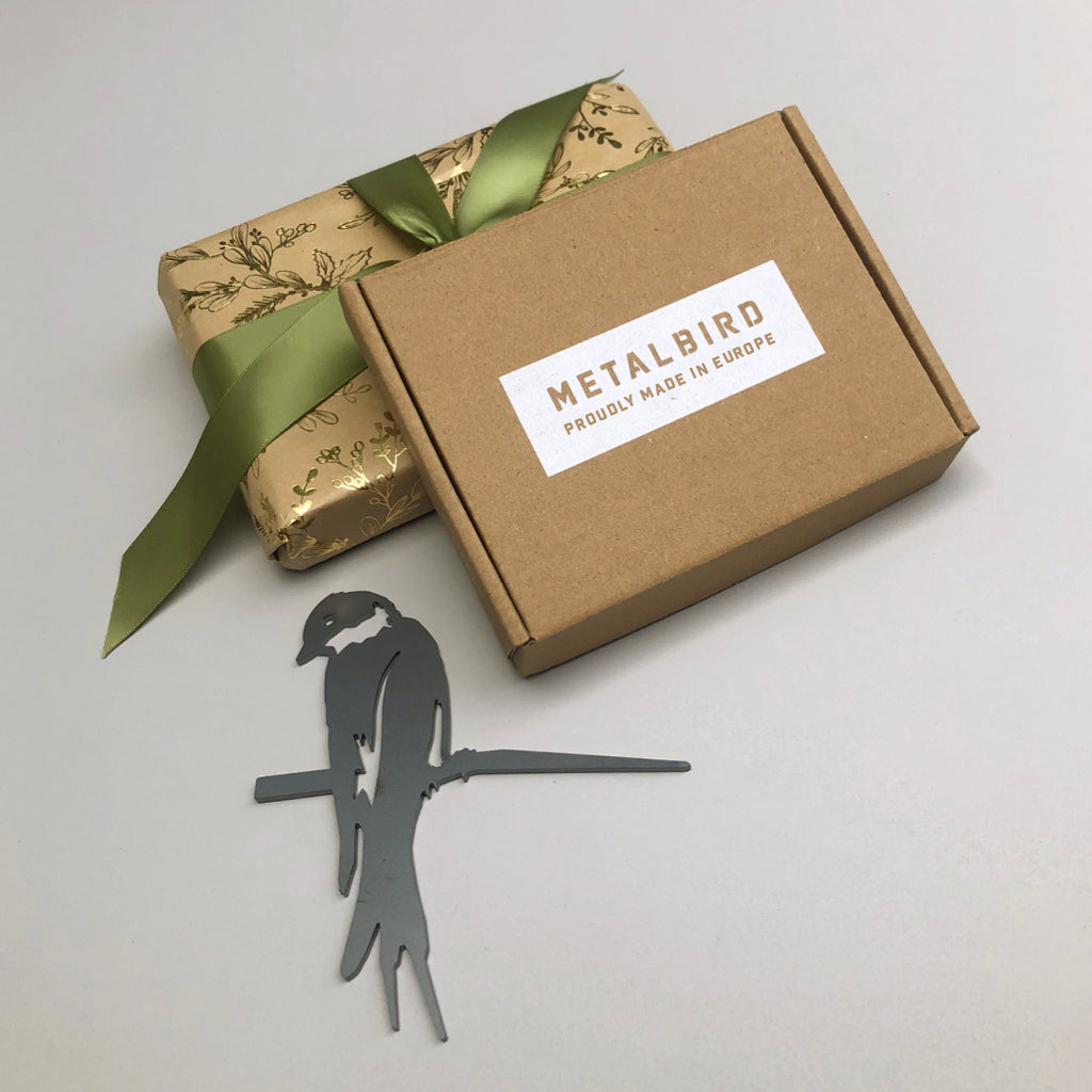 A small cardboard box labeled Metalbird UK sits next to a gold gift wrapped with a green ribbon. In front, the Baby Bird Bundle - Swallow Trio, corten steel silhouettes of birds on branches, lies flat—perfect for unique garden art.