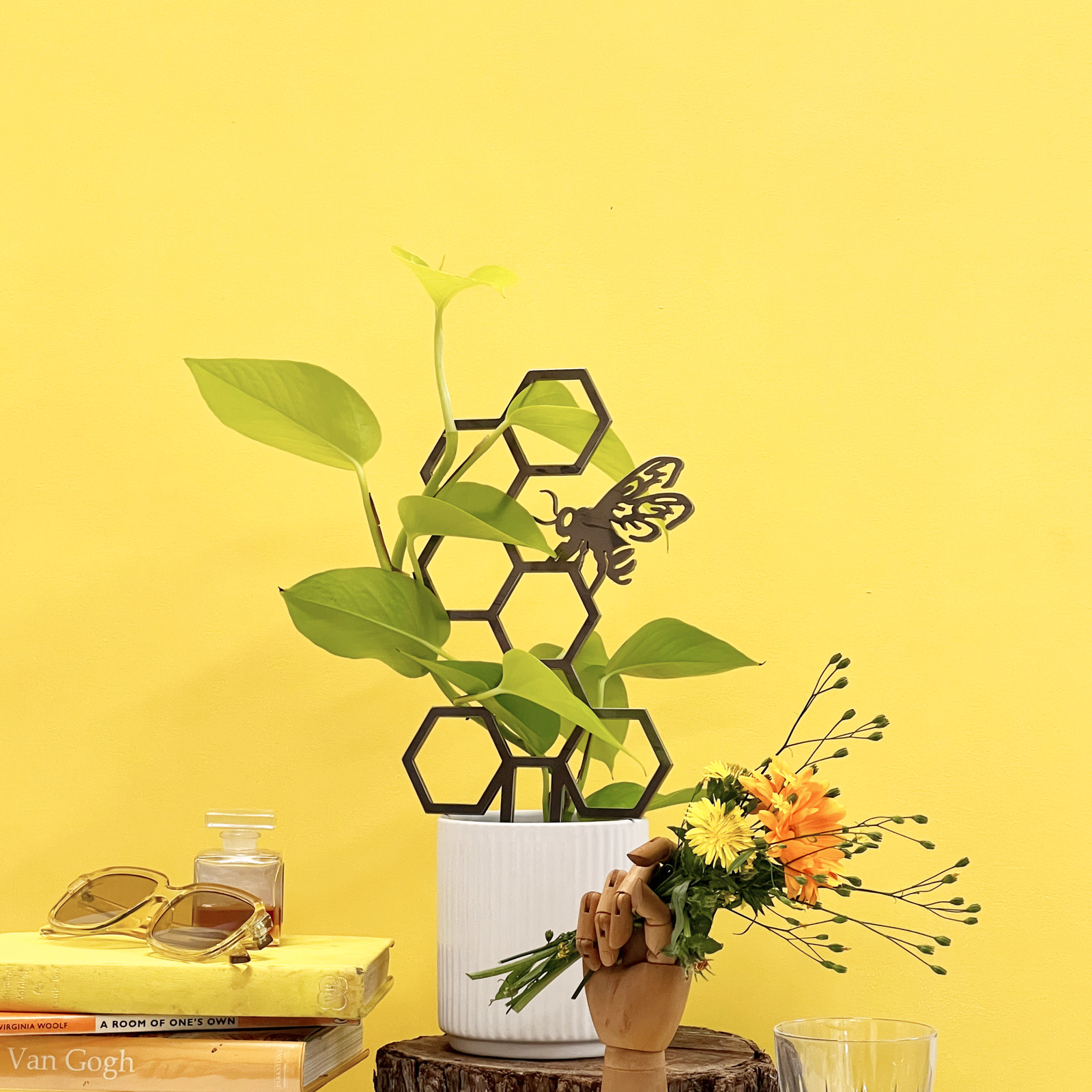 The Metalbird UK Honeybee Plant Trellis, a honeycomb-shaped metal trellis in a white pot with green plants and butterfly garden art, sits on a yellow table surrounded by books, sunglasses, a glass, and a wooden hand holding yellow flowers.