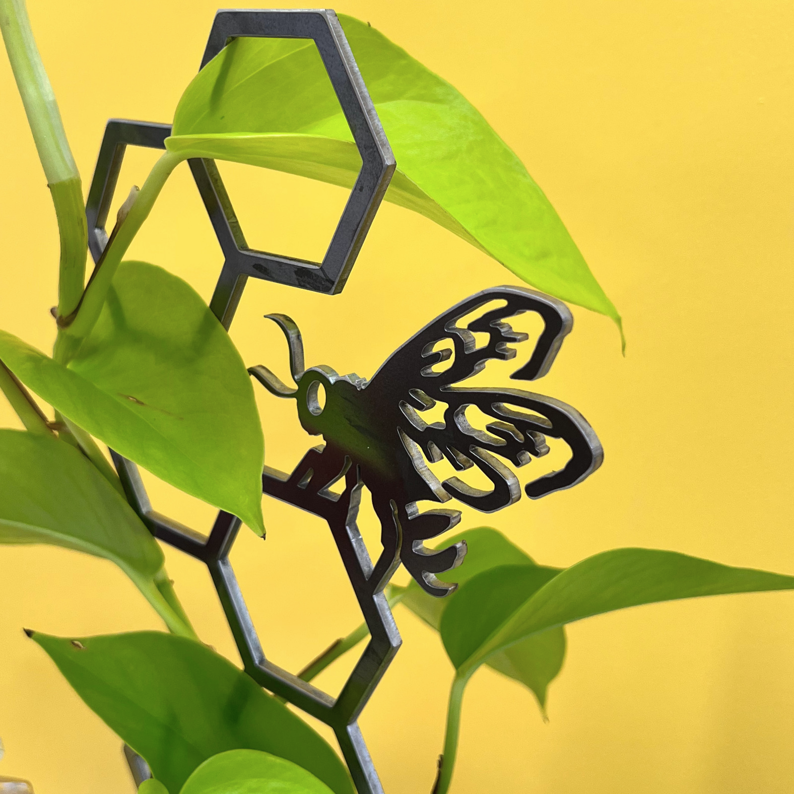 The Honeybee Plant Trellis by Metalbird UK features a black metal bee and honeycomb design, set among green leaves with a yellow backdrop—a unique gift idea for garden lovers.