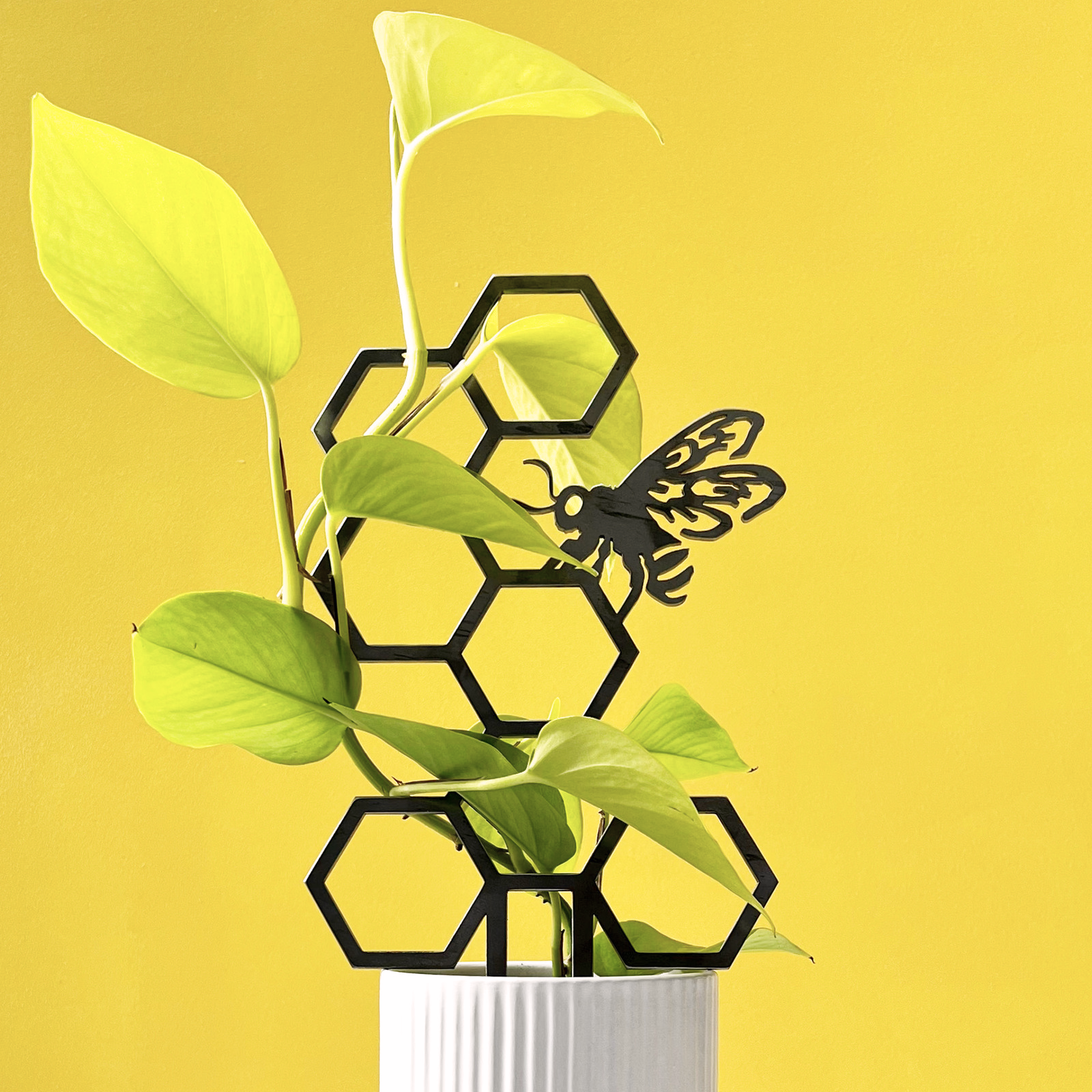 The Metalbird UK Honeybee Plant Trellis features green leaves climbing through a black corten steel honeycomb and bee design, set in a white ribbed pot with a bright yellow backdrop—an eye-catching garden accent.