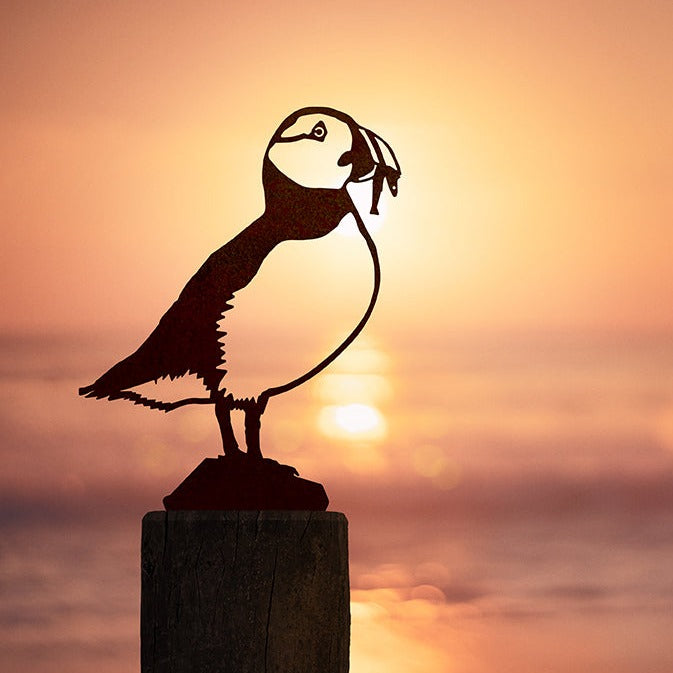 The Metalbird UK Puffin, a metal garden silhouette, is displayed on a wooden post at sunset by calm water, with the sun’s reflection shimmering on the surface.