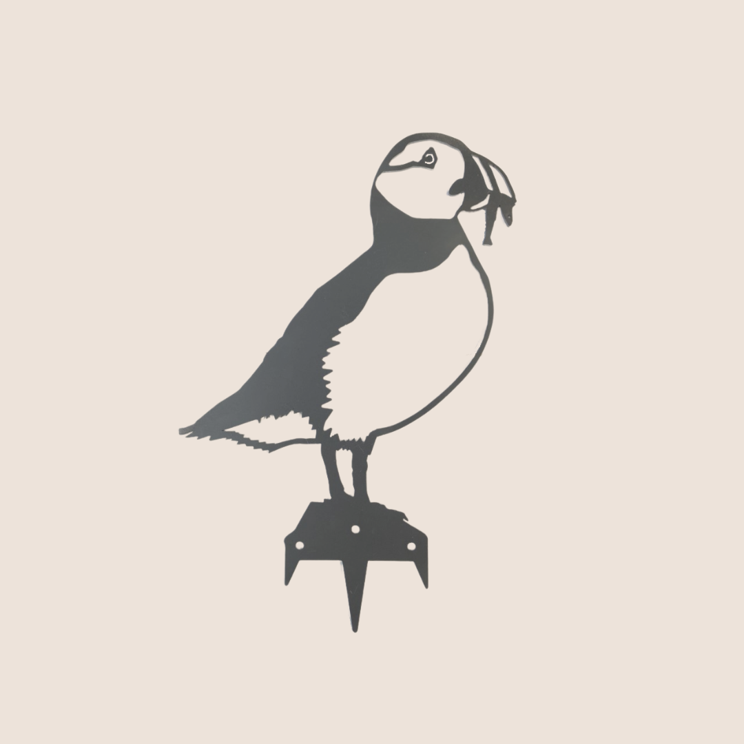 The Metalbird UK Puffin features a minimalist black silhouette inspired by corten steel art, displayed on a small platform against a plain light beige background.