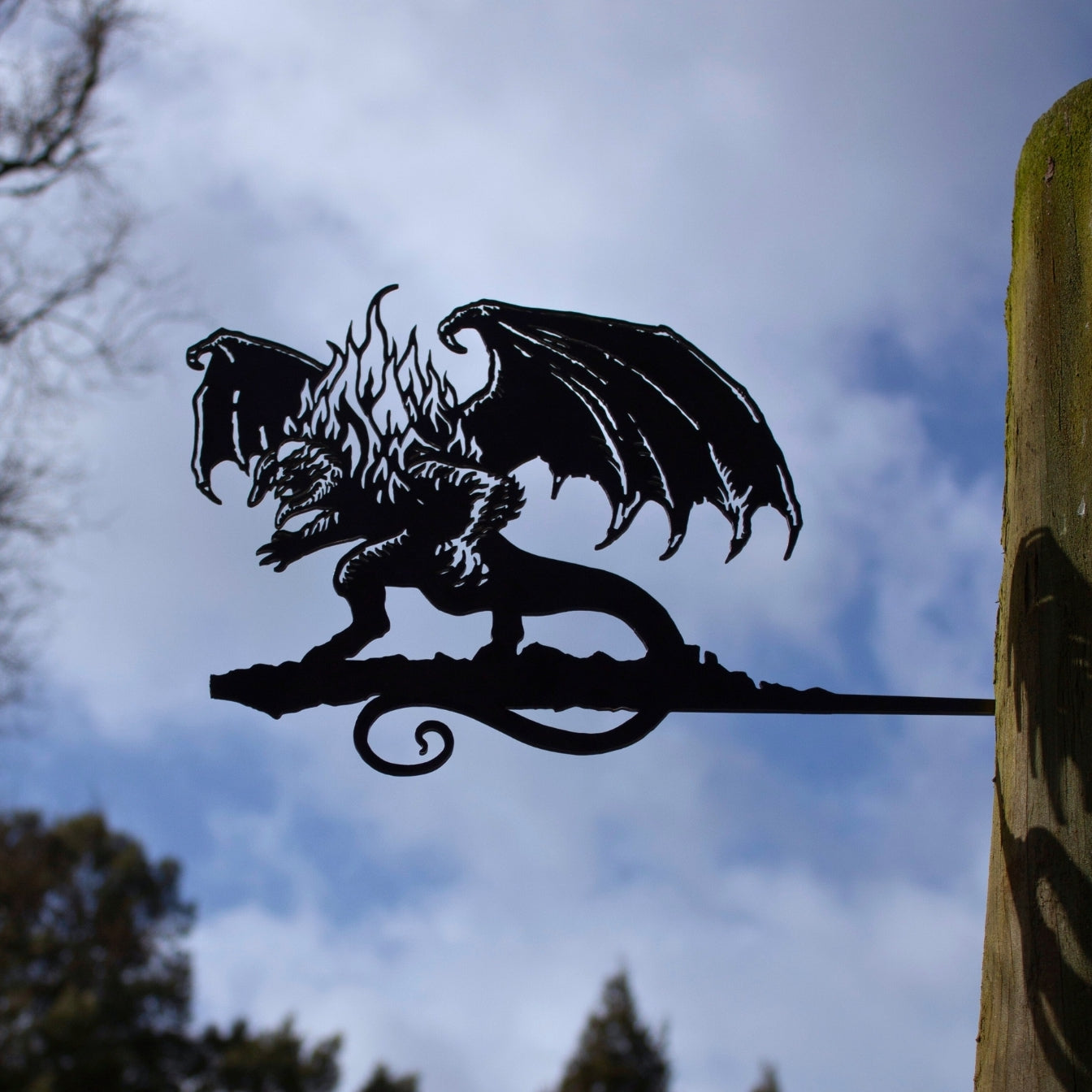 A striking corten steel BALROG™ silhouette by Metalbird UK, featuring wings and flames, is mounted on a wooden post against a cloudy sky with tree branches—an unforgettable gift for any fantasy lover.