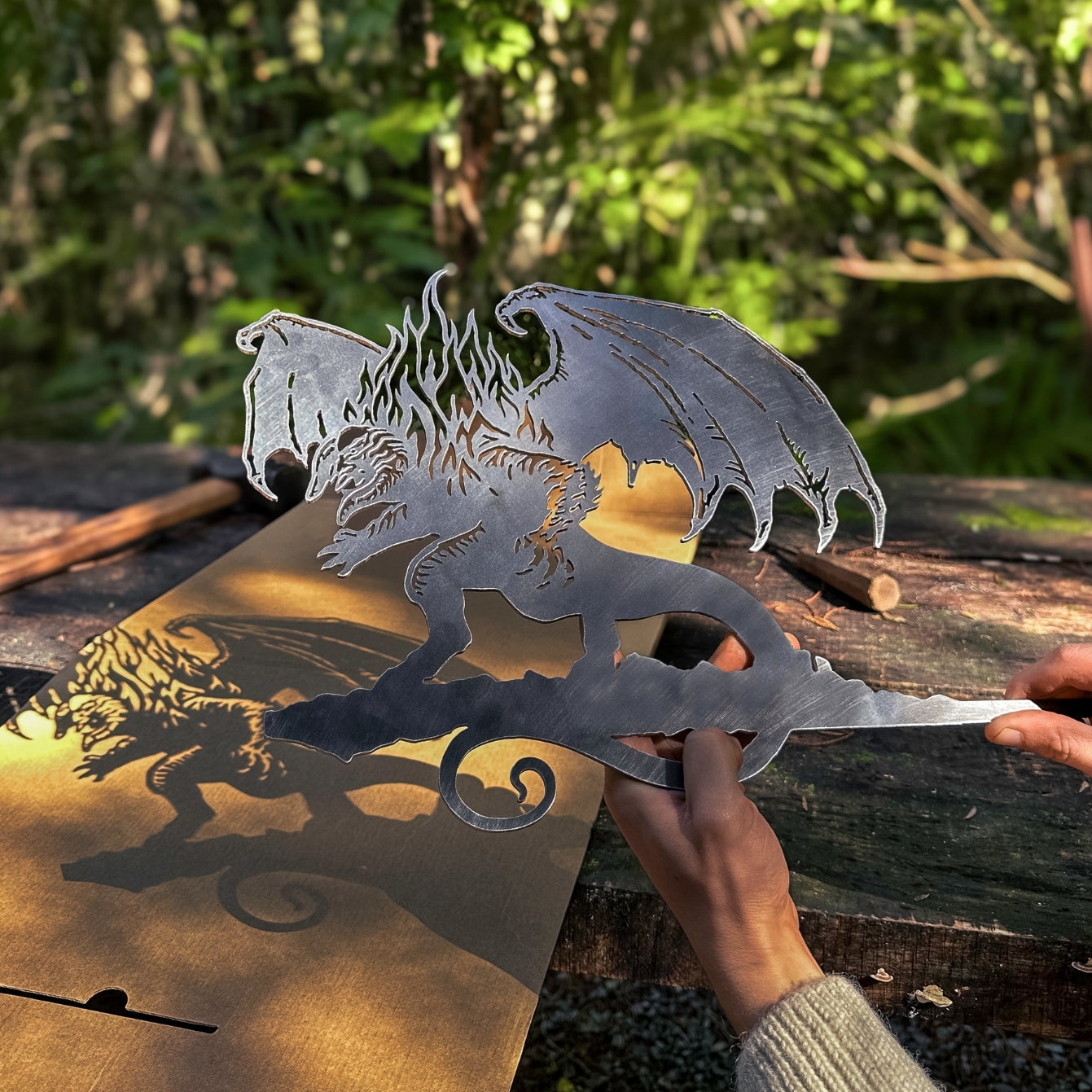 A person holds the BALROG™ by Metalbird UK—a premium metal dragon perched on a branch—casting a detailed shadow outdoors amid lush greenery, making it a striking piece of yard art.
