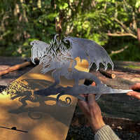 A person holds the BALROG™ by Metalbird UK—a premium metal dragon perched on a branch—casting a detailed shadow outdoors amid lush greenery, making it a striking piece of yard art.
