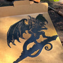 The BALROG™ by Metalbird UK is a black dragon silhouette with large wings and a curled tail, displayed on brown cardboard against wood. Sunlight reveals the premium yard art details of this striking metal figure.