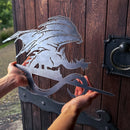 A person holds the FELL BEAST™ corten steel sign by Metalbird UK, shaped like a dragon on a curved staff, standing before a dark wooden door with medieval wrought iron hinges and a round handle.