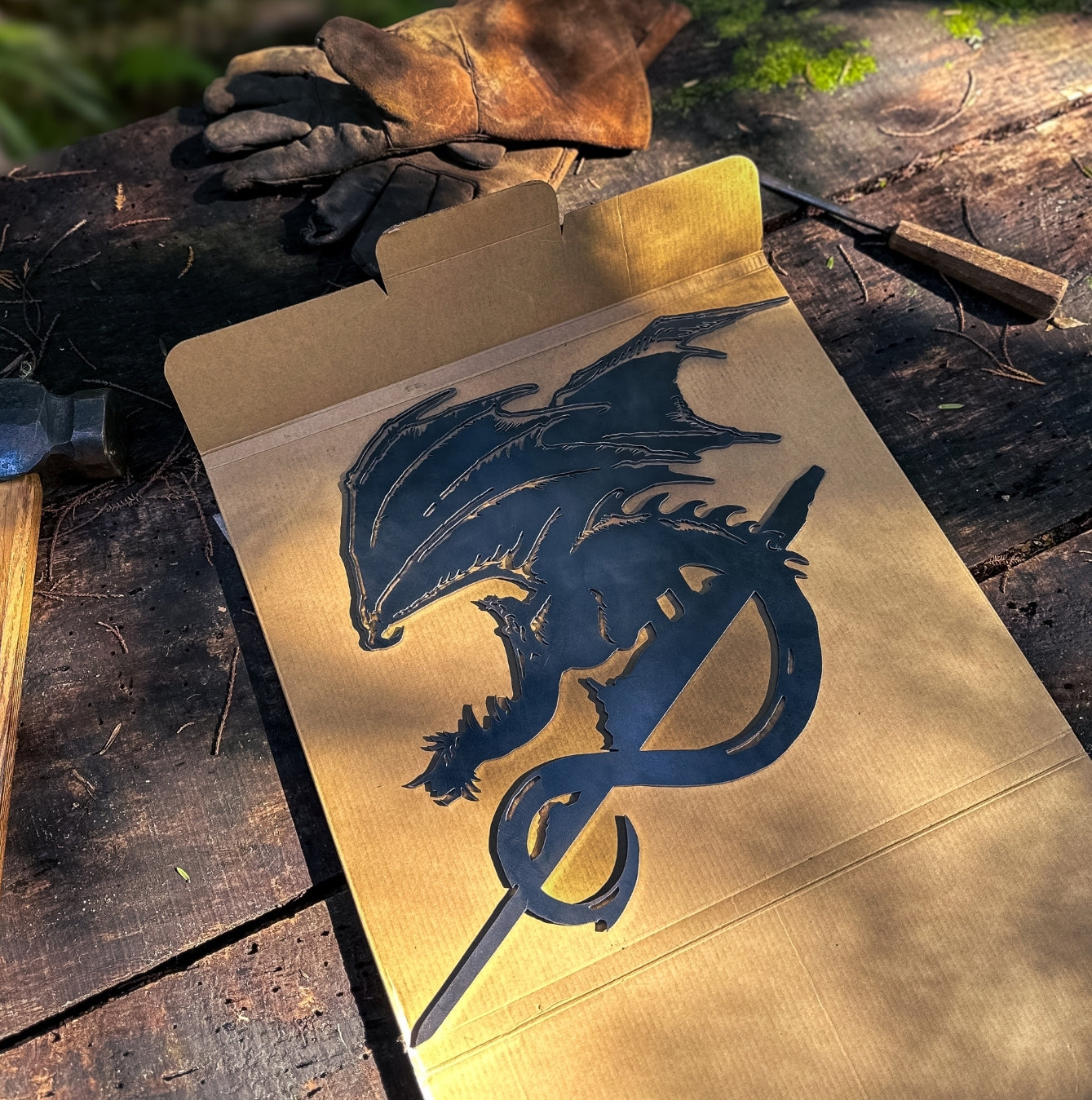 The FELL BEAST™ by Metalbird UK—a premium black metal dragon silhouette resting on a cardboard sheet atop a rustic wooden table with work gloves, hammer, and chisel in sunlight—makes a unique gift for any metal art enthusiast.