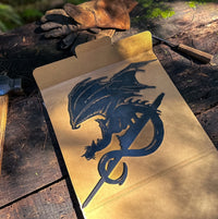 The FELL BEAST™ by Metalbird UK—a premium black metal dragon silhouette resting on a cardboard sheet atop a rustic wooden table with work gloves, hammer, and chisel in sunlight—makes a unique gift for any metal art enthusiast.