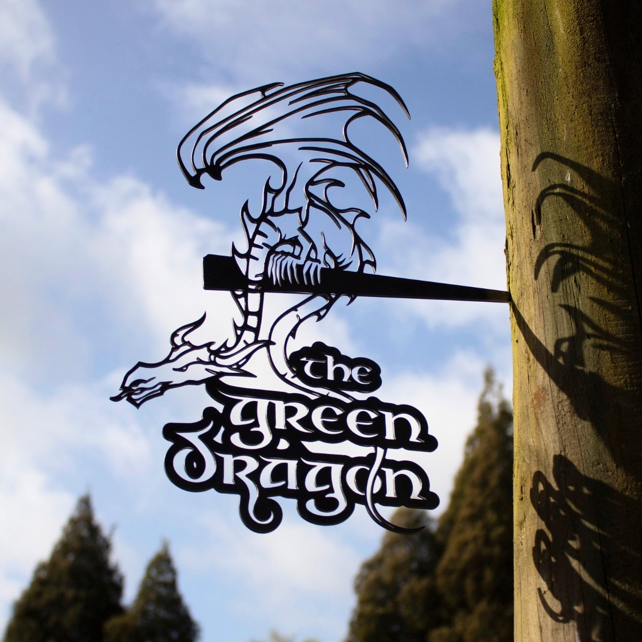 THE GREEN DRAGON™ by Metalbird UK: A corten steel dragon sign hangs from a wooden post, its silhouette bold against trees and a blue sky. An ideal gift for any fantasy fan.