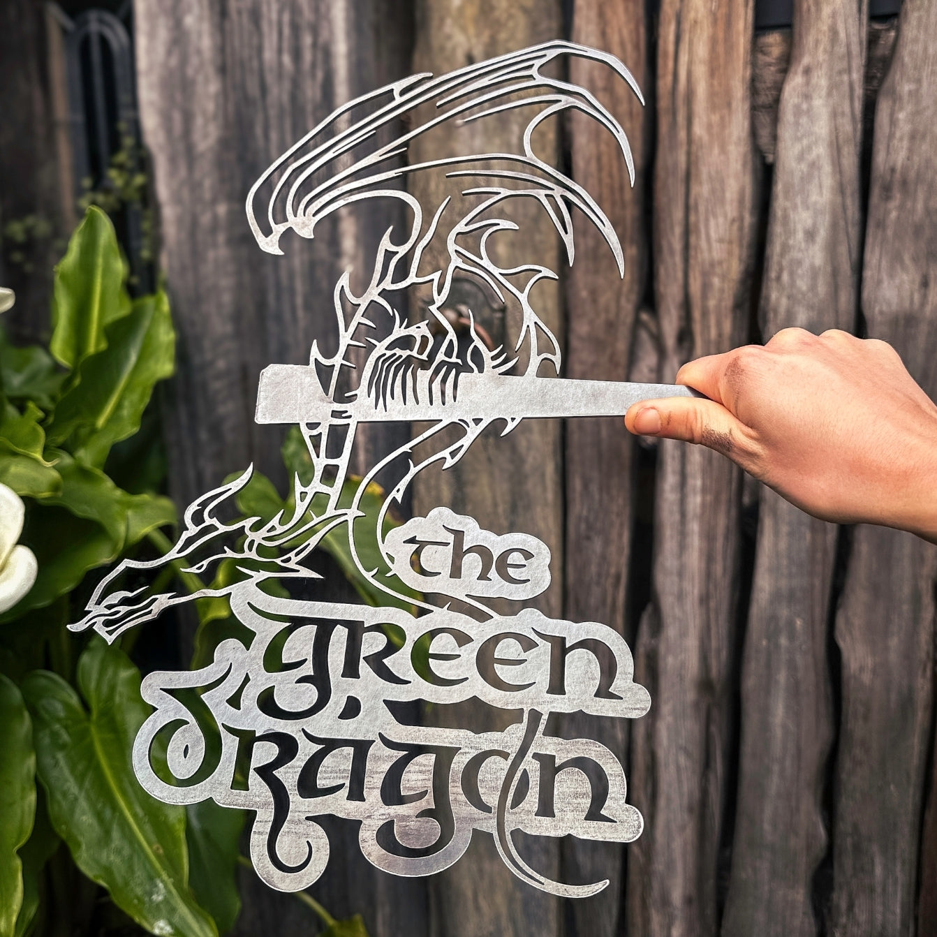 A hand holds a premium metallic sign by Metalbird UK featuring a stylized dragon above "THE GREEN DRAGON™" in decorative lettering, set against a wooden fence and green foliage.