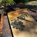 A cardboard box featuring THE GREEN DRAGON™ by Metalbird UK, with a dragon-and-sword silhouette design, sits on a rustic outdoor wooden surface—sunlight, gloves, and wood nearby inspire creative yard art.