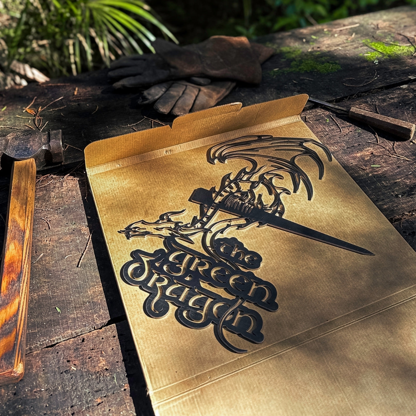 A cardboard box featuring THE GREEN DRAGON™ by Metalbird UK, with a dragon-and-sword silhouette design, sits on a rustic outdoor wooden surface—sunlight, gloves, and wood nearby inspire creative yard art.