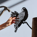 A hand holds the Manumea black metal bird silhouette by Metalbird UK - CC, perched on a branch with a hammer nearby and a tree stump on the right—unique inspiration for creative yard art.