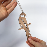 A person holds a Metalbird UK Wooden Hanging Decoration shaped like a bird on a branch, suspended by a white string against a plain background—an ideal gift for nature lovers.