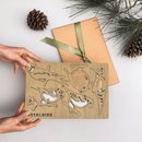 A person holds a set of premium Wooden Hanging Decorations by Metalbird UK next to a brown paper gift with a green ribbon, pinecone, and pine branches—perfect for nature lovers’ yard art.