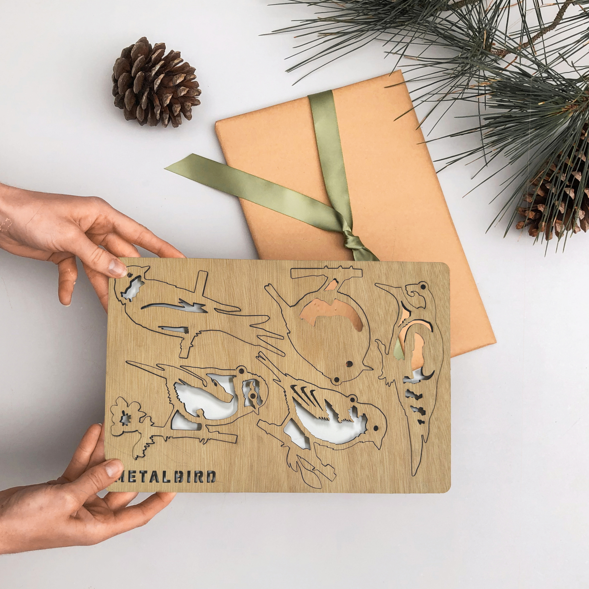 A person holds a set of premium Wooden Hanging Decorations by Metalbird UK next to a brown paper gift with a green ribbon, pinecone, and pine branches—perfect for nature lovers’ yard art.