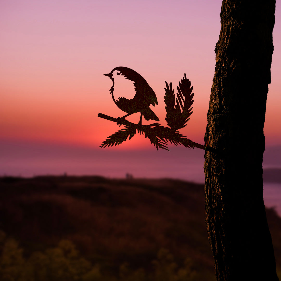 The Metalbird UK Robin Redbreast is a Corten steel bird ornament perched on a branch, mounted to a tree with a blurred landscape and sunset backdrop—an ideal Valentine's Day gift.