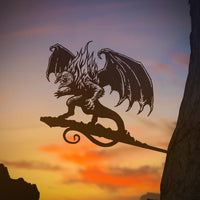 The BALROG™ by Metalbird UK is a bold metal yard art piece featuring a winged, horned silhouette. Mounted on a tree, it stands out against a vivid sunset sky of orange, pink, and purple.