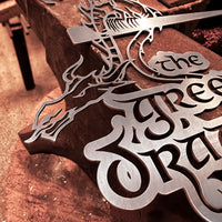 A close-up of THE GREEN DRAGON™ corten steel sign by Metalbird UK, showcasing an intricate dragon silhouette and crafted text, displayed on a wooden workbench—a striking gift for any art lover.