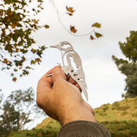 A hand holds up the Mini Woodpecker silhouette by Metalbird UK in an outdoor setting, with trees, a grassy hillside, and a partly cloudy sky visible in the background.