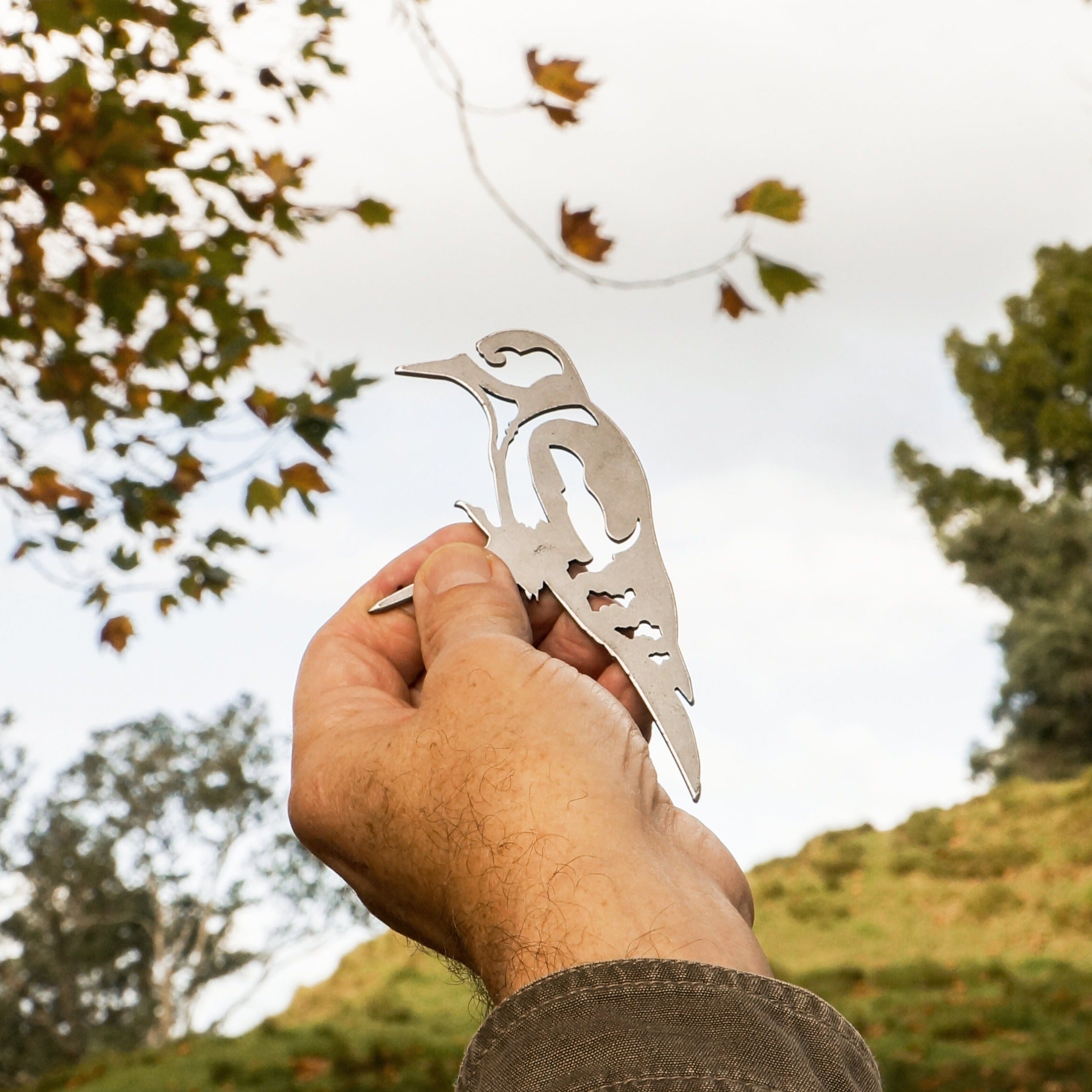 A hand holds up the Mini Woodpecker silhouette by Metalbird UK in an outdoor setting, with trees, a grassy hillside, and a partly cloudy sky visible in the background.