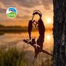 Silhouette of an Osprey perched on a tree at sunset by a lake, perfect as yard art or a unique gift from Metalbird UK. Grassy reeds and blurred water create a tranquil backdrop. The RSPB logo and ‘Proud to partner with’ are shown in the top left corner.