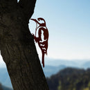 A tree trunk in the foreground features the Great Spotted Woodpecker - Seconds by Metalbird UK, a premium corten steel silhouette; mountains and blue sky create a blurred background.