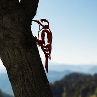 A tree trunk in the foreground features the Great Spotted Woodpecker - Seconds by Metalbird UK, a premium corten steel silhouette; mountains and blue sky create a blurred background.