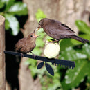 A small brown chick and adult bird perch together on the Metalbird UK Stick Feeder, a corten steel silhouette sign, creating charming yard art with blurred green foliage in the background.