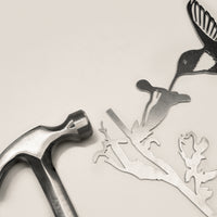 A metal hammer rests beside intricately cut corten steel shapes—a hummingbird, a flower, and branches—on a light surface. The "Hummingbird" by Metalbird UK makes an inspiring gift for metal art enthusiasts.