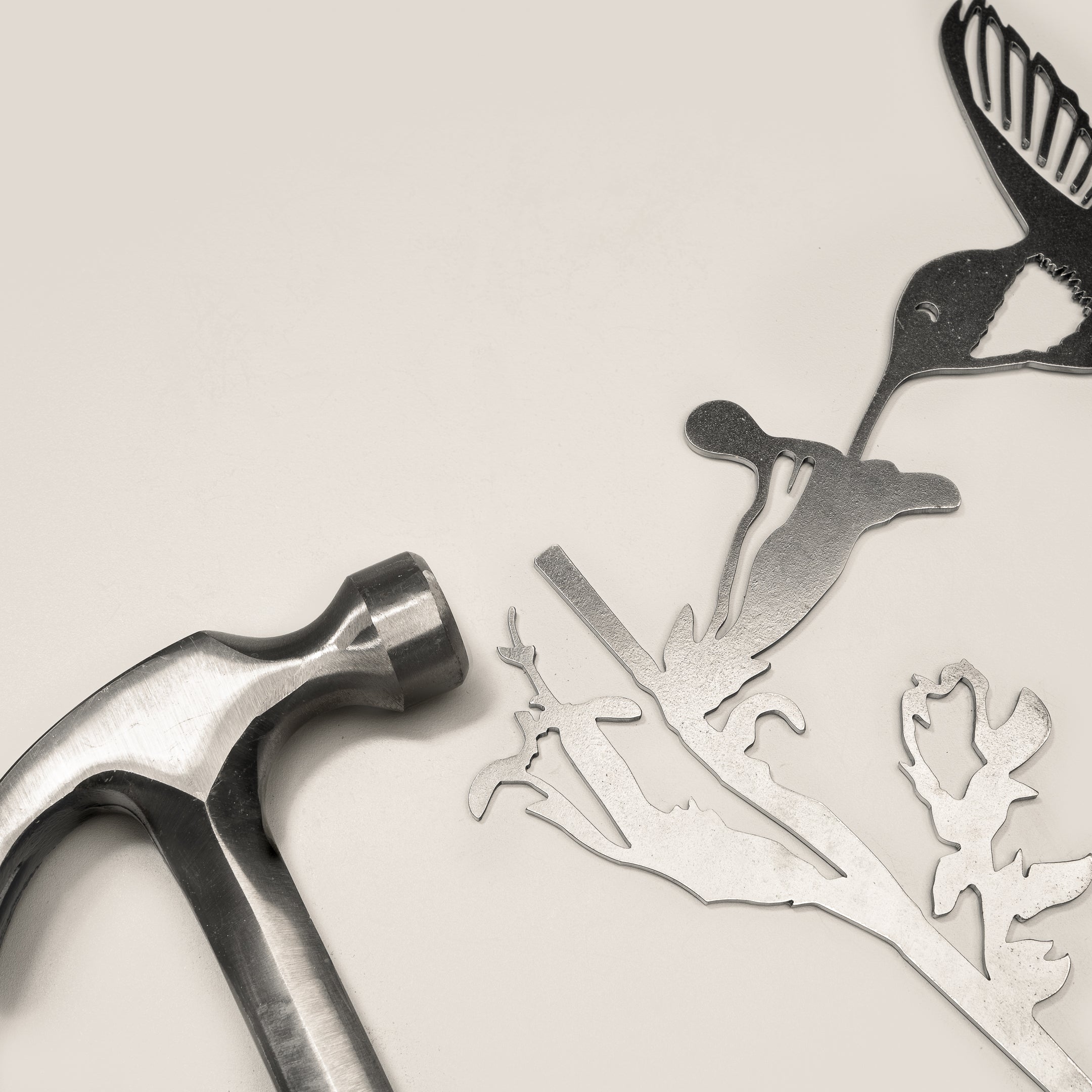 A metal hammer rests beside intricately cut corten steel shapes—a hummingbird, a flower, and branches—on a light surface. The "Hummingbird" by Metalbird UK makes an inspiring gift for metal art enthusiasts.
