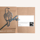 The Turtle Dove by Metalbird UK is a metal bird silhouette sculpture perched on a branch, displayed atop a brown cardboard folder with recycling icons, a printed message, and a black-and-white photo inside.