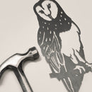 A silver hammer lies next to the Metalbird UK Barn Owl, a detailed corten steel owl silhouette with unique feather patterns, both arranged on a light beige surface.