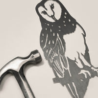 A silver hammer lies next to the Metalbird UK Barn Owl, a detailed corten steel owl silhouette with unique feather patterns, both arranged on a light beige surface.